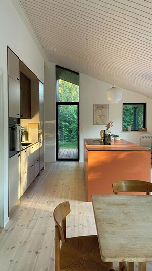 A modern kitchen with a large window, wooden flooring, and a dining area with a table and chairs. - Beautiful cottage with a simple modern aesthetic (Vejby Strand)
