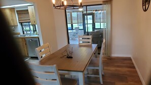 A dining area with a wooden table and white chairs, a kitchen with wooden cabinets, and a view of the outdoors through a glass door. - Bicycles, fishing poles and lap tops invited! (Inverness)