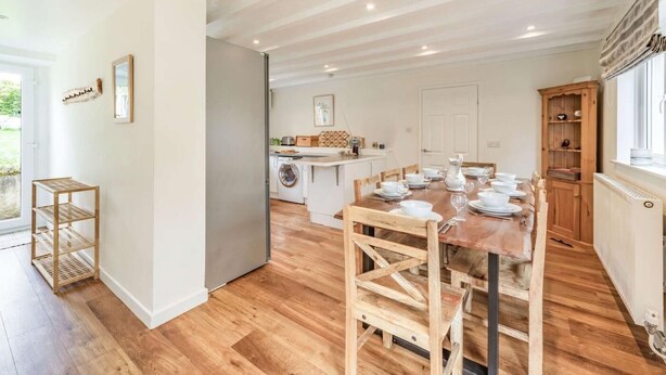 Un'ampia sala da pranzo con un tavolo di legno apparecchiato per quattro persone, una cucina sullo sfondo e un'ampia finestra a destra.