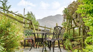 A stone patio with metal chairs and a table, surrounded by greenery and a wooden fence. - 2 Bed in Keswick (oc-sz302) (Rosthwaite)