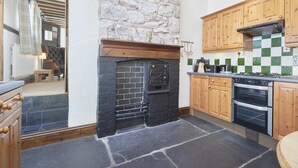 A kitchen with stone walls, wooden cabinets, a black fireplace, and a tiled backsplash. - 1 Bed in Holmrook (oc-sz471) (Irton with Santon)