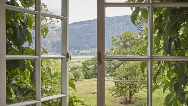 Blick durch ein fensterförmiges Gitter, das eine Landschaft mit Bäumen und einer Hügelkette in der Ferne zeigt.
