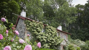 Ein Steinhäuschen mit rotem Dach, umgeben von üppiger Vegetation und blühenden Pflanzen.