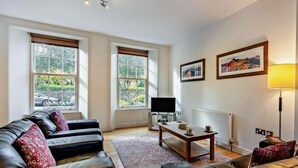 Una sala de estar con un sofá de cuero, una mesa de centro, un televisor y ventanas amplias con vistas a zonas verdes.