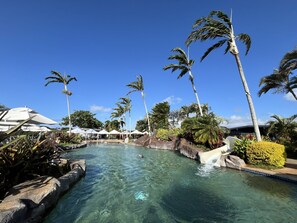 Ein Pool mit klarem Wasser, Palmen, einem Wasserfall und Sitzmöglichkeiten im Freien mit Sonnenschirmen.
