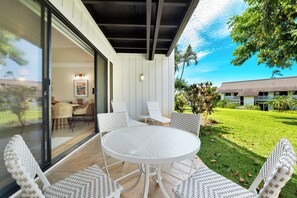 A covered outdoor seating area with a round table and wicker chairs, overlooking a well-maintained lawn and a building in the background. - Luxe Kauai 1BR Retreat | A/C & Beach Access (Koloa)