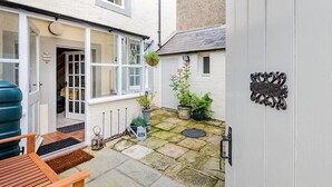 Ein weißes Gebäude mit einer Tür, auf der 'WELCOME' steht, ein kleiner Garten mit Topfpflanzen und eine hölzerne Bank.