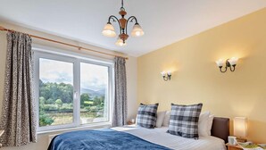 A bedroom with a large bed, a window with curtains, a chandelier, wall sconces, and a nightstand with a lamp. - 3 Bed in Keswick (oc-sz249) (Keswick)