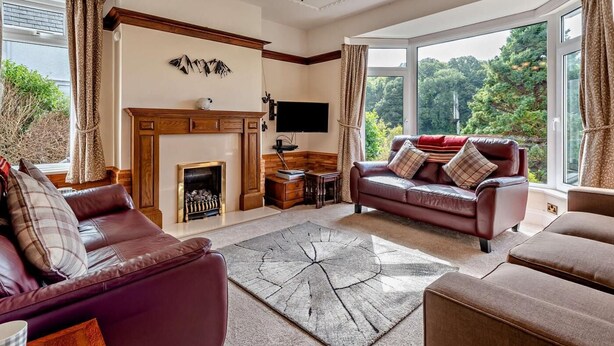 A living room with a fireplace, leather sofas, a chandelier, and a large window with a view of greenery. - 3 Bed in Keswick (oc-sz249) (Keswick)