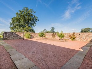 A courtyard with a gravel path, stone walls, and a large tree. - 4 Bed in Penrith (oc-sz383) (Skirwith)