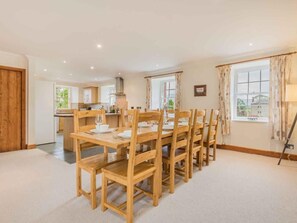 A spacious dining area with wooden tables and chairs, a kitchen in the background, and large windows with curtains. - 4 Bed in Penrith (oc-sz383) (Skirwith)