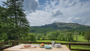 A wooden picnic table with a croissant, plates, and cups, overlooking a green landscape with hills. - 3 Bed in Broughton-in-furness (oc-sz107) (Eskdale)