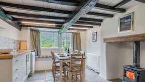 A rustic kitchen with a stone floor, wooden ceiling beams, a dining table with chairs, a stove, and a window with curtains. - 3 Bed in Broughton-in-furness (oc-sz107) (Eskdale)