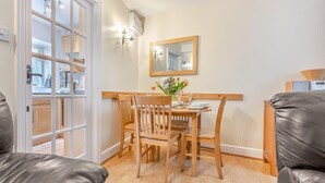 Un coin repas avec une table en bois, quatre chaises, un vase de fleurs, un miroir au mur et une vue sur la cuisine par une porte vitrée.