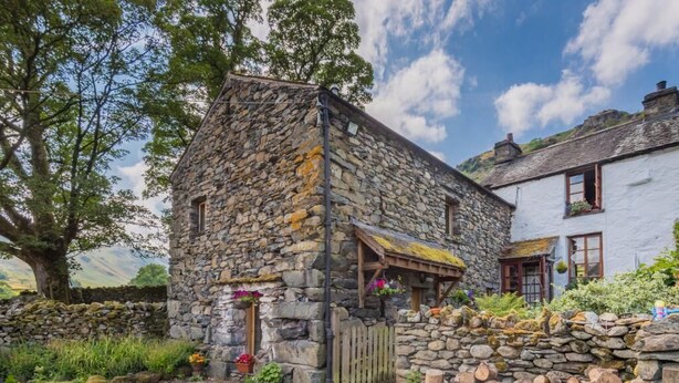 A stone building with a wooden door, a stone wall, and a garden with potted plants. - 2 Bed in Penrith (oc-sz470) (Patterdale)