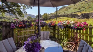 A patio with a table and chairs, a hanging flower basket, and a stone wall in the background. - 2 Bed in Penrith (oc-sz470) (Patterdale)