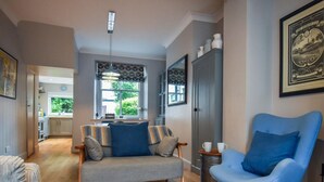 A living room with a sofa, a blue armchair, a dining table, and a kitchen in the background. - 3 Bed in Keswick (oc-sz029) (Keswick)