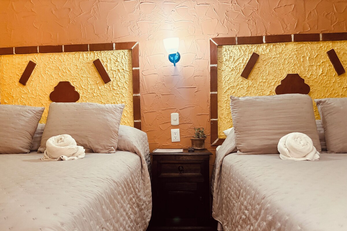 A hotel room with two beds, each with a pillow and a rolled towel. There is a nightstand with a lamp and a small plant.