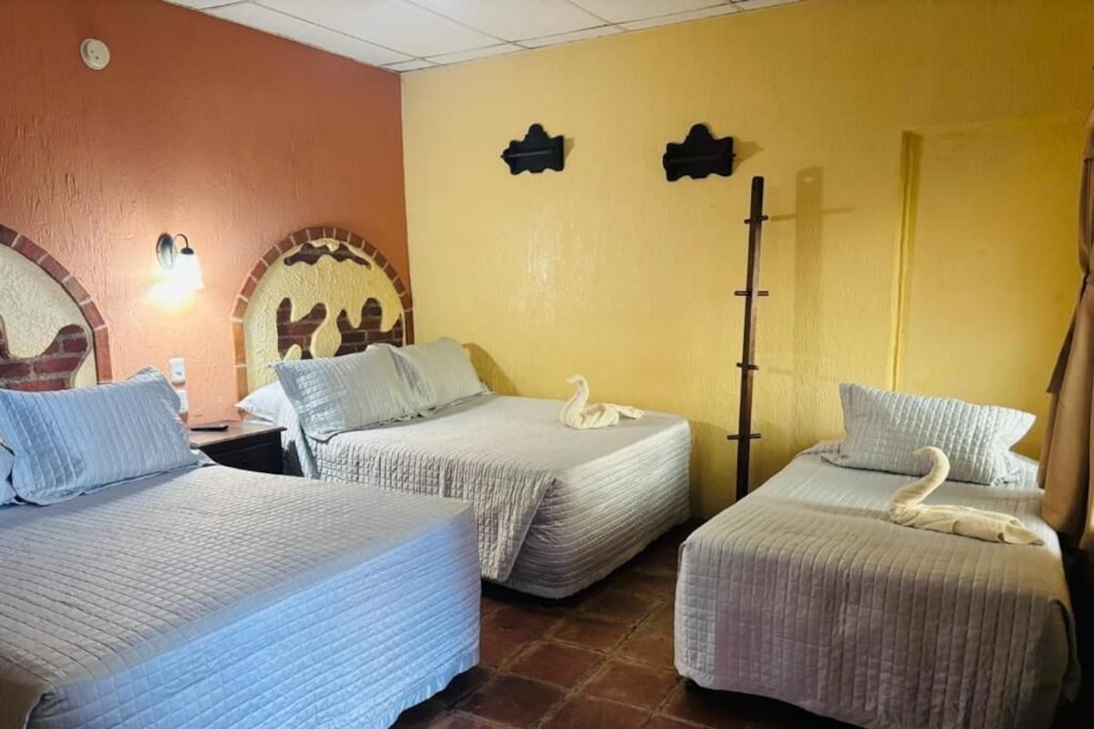 A hotel room with three beds, each with a swan-shaped pillow, a wall-mounted light fixture, and a door with a vertical handle.