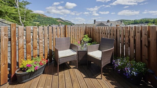 A wooden deck with wicker chairs and a table, surrounded by a wooden fence, with a view of hills and a clear sky. - 1 Bed in Keswick (oc-sz166) (Keswick)
