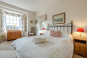 Ein Schlafzimmer mit einem Bett, einem Holzkleiderschrank, einem Sessel, einem Nachttisch und einem Fenster mit Vorhängen.