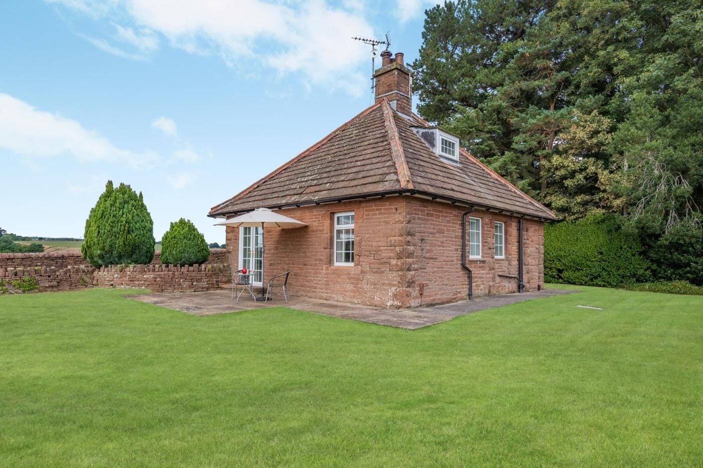A brick house with a sloped roof, a small porch with a table and chairs, and a well-maintained lawn.