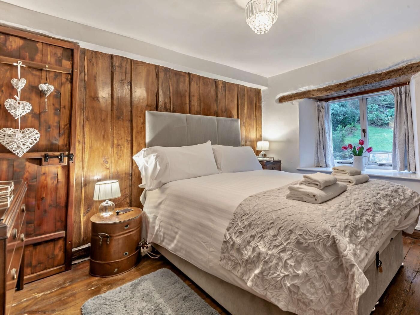 A bedroom with a wooden wall, a bed with white linens, a nightstand, a lamp, and a window with a view of greenery.
