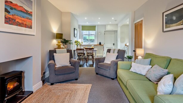 Ein Wohnzimmer mit Kamin, zwei Sesseln, einem grünen Sofa und einer Küche im Hintergrund.