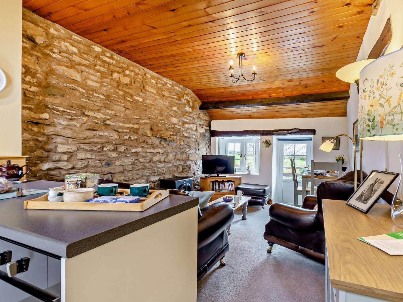 A cozy interior with a stone wall, wooden ceiling, a dining area with a table and chairs, and a living room with a sofa and a television.