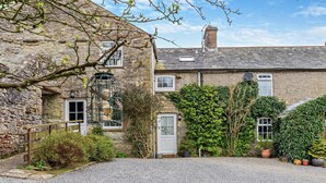 A stone building with a gravel driveway, a white door, and a small garden with potted plants. - 1 Bed in Kirkby Stephen (oc-sz355) (Newbiggin-on-Lune)