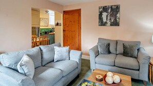 Una sala de estar con dos sofás grises, una mesa de centro de madera con objetos decorativos, una puerta de madera y una cocina visible a través de una puerta abierta.