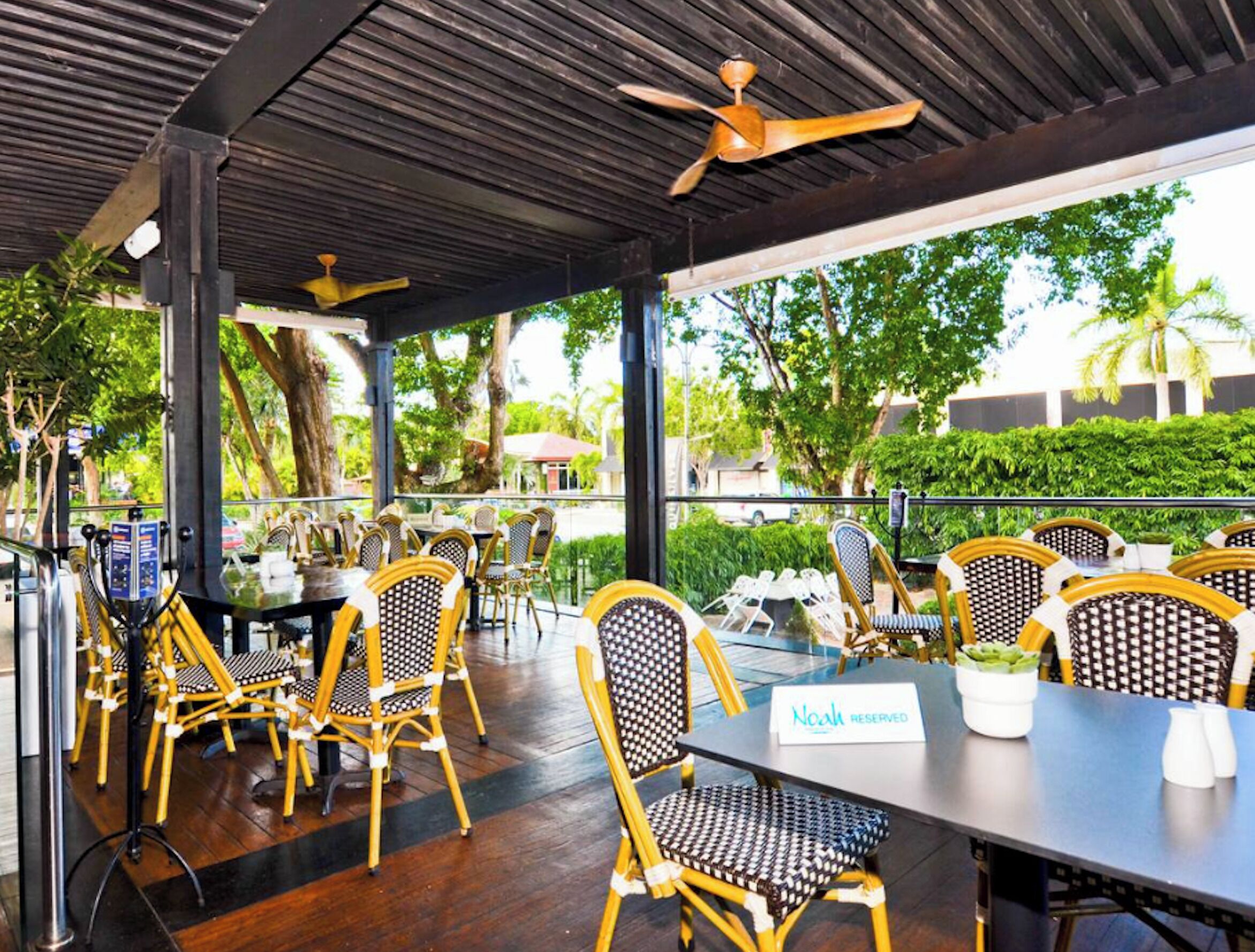 Un restaurant en plein air avec des chaises et des tables en osier, un ventilateur de plafond et une vue sur la verdure et les bâtiments.