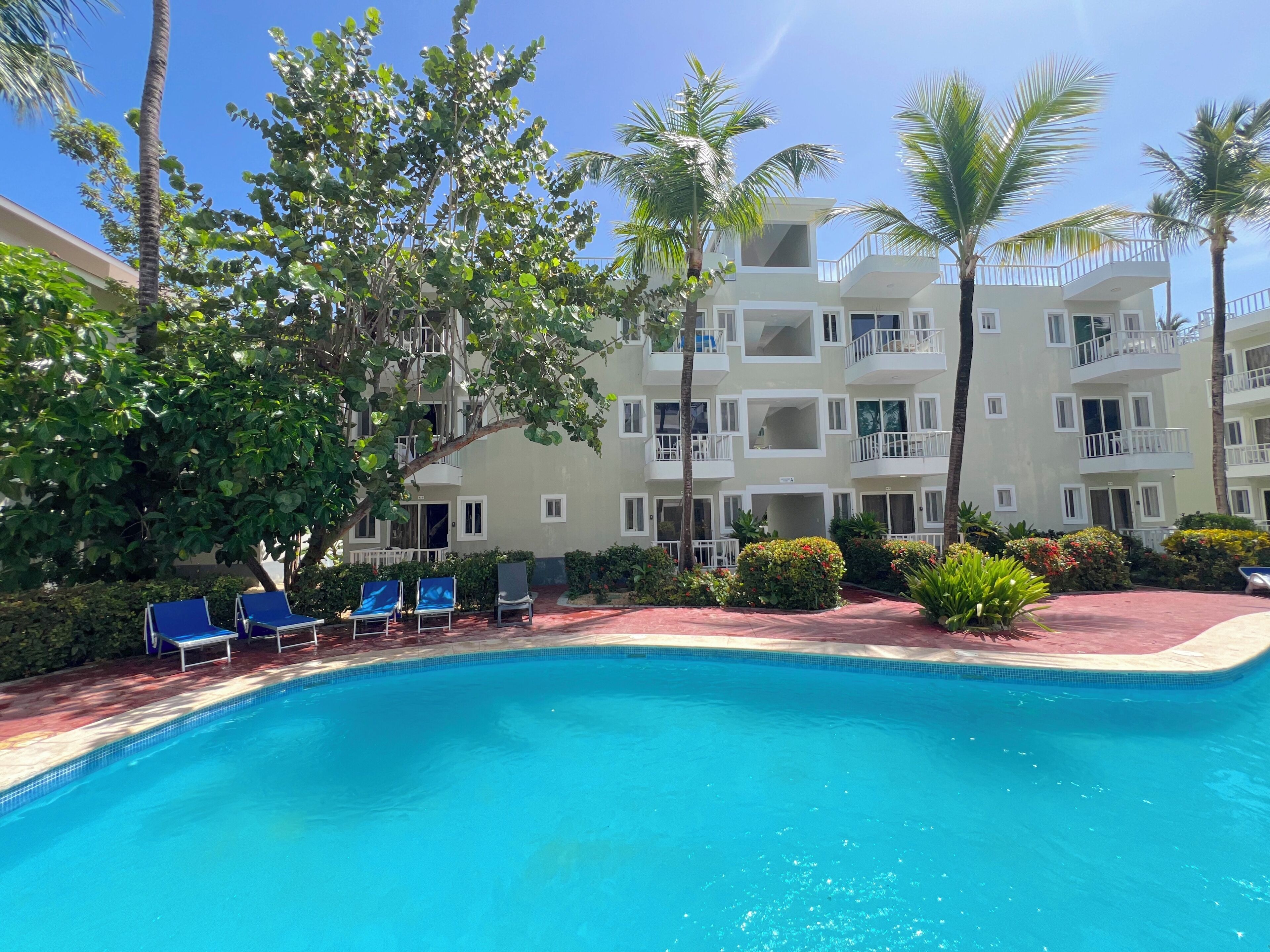 A pool area with lounge chairs, a building with balconies, and palm trees.