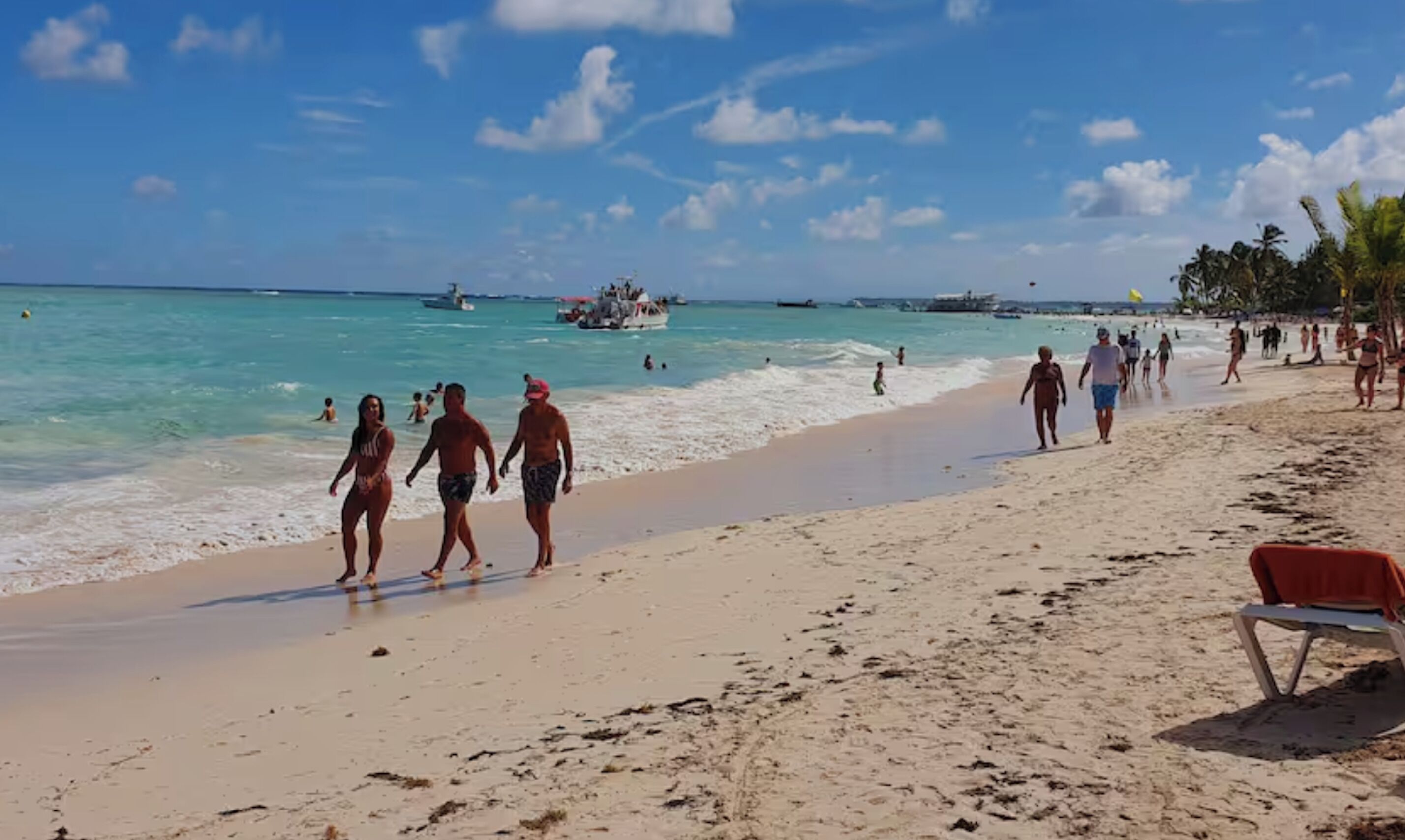 Uma praia com pessoas caminhando pela margem, barcos na água e um céu claro.