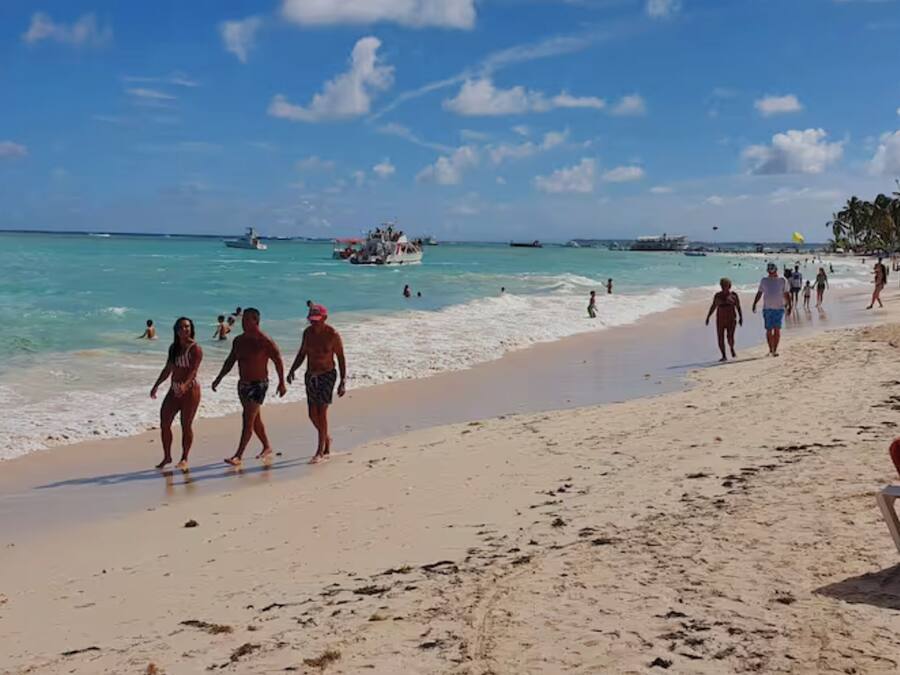 Uma praia com pessoas caminhando pela margem, barcos na água e um céu claro.