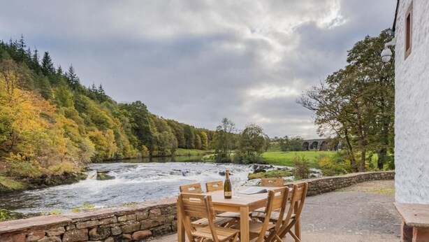 Un espace extérieur aménagé au bord de la rivière, avec une table et des chaises, un mur en pierre et un bâtiment en arrière-plan.