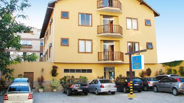 A multi-story yellow building with a balcony on the second floor, a parking lot with several cars, and a sign indicating a hotel. - Village du Soleil (Cabo Frio)
