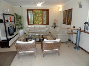 A living room with a sofa, two wicker chairs, a coffee table, a television, a bookshelf, a window with a view, and a wall-mounted lamp.