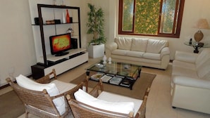 A living room with a sofa, two wicker chairs, a coffee table, a TV, a bookshelf, and a window with a view. - Village du Soleil (Cabo Frio)
