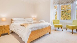 Un dormitorio con cama de madera, mesitas de noche, ventana con vistas a árboles y dos sillas amarillas.
