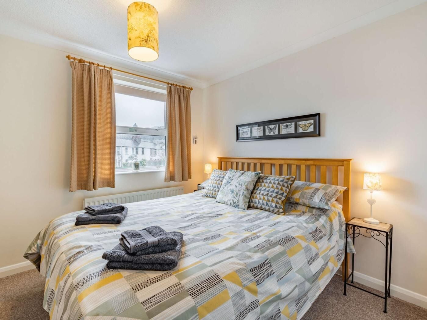 A bedroom with a bed, a window with curtains, a bedside table with a lamp, and a wall-mounted picture frame.