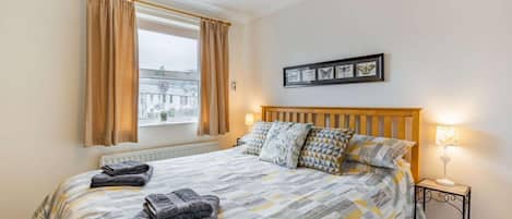 A bedroom with a bed, a window with curtains, a bedside table with a lamp, and a wall-mounted picture frame.