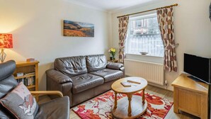 Ein Wohnzimmer mit einem Ledersofa, einem runden Couchtisch aus Holz, einem Bücherregal, einem Flachbildfernseher und einem Fenster mit Vorhängen.