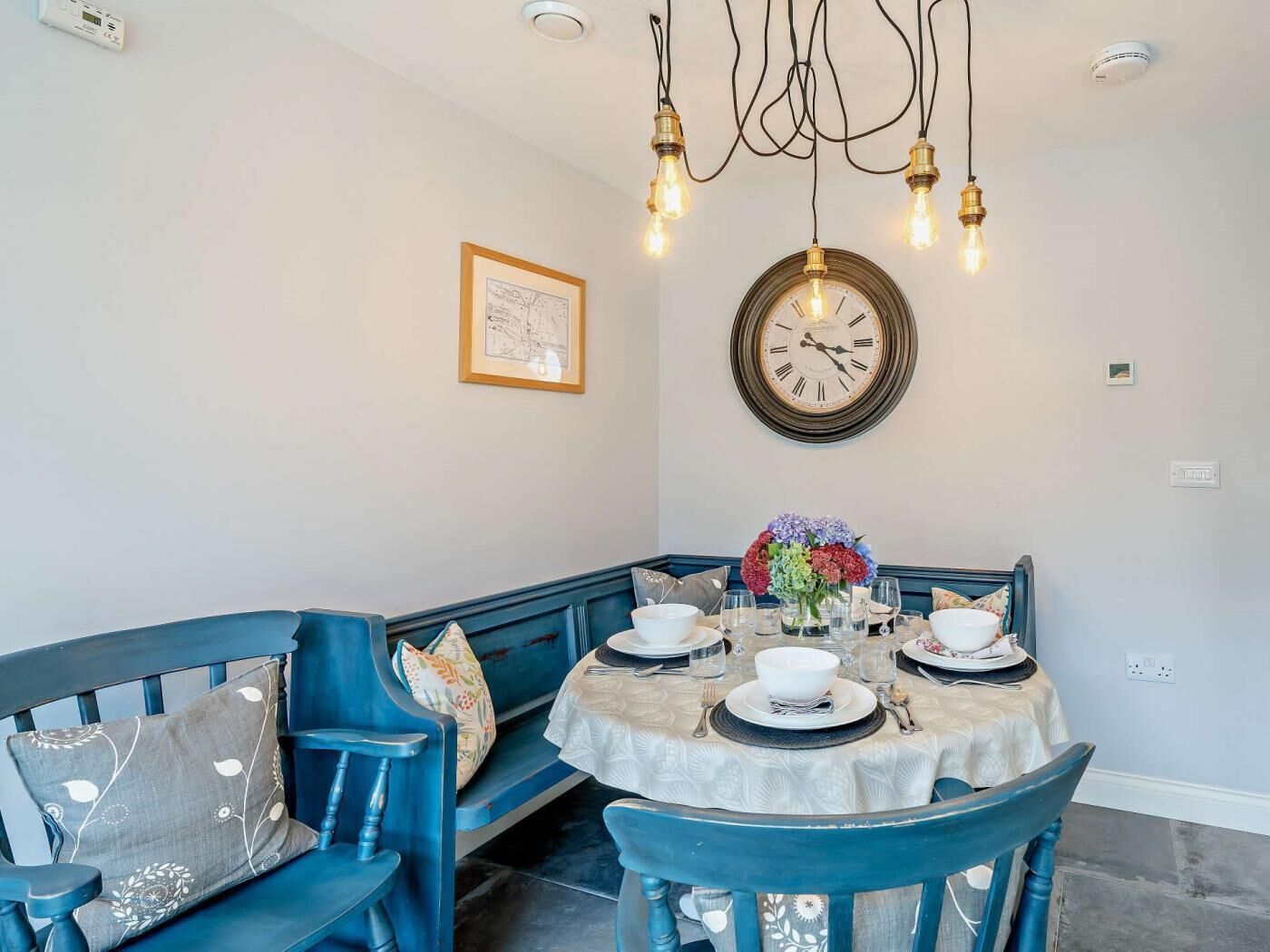 A dining area with a round table set for four, a blue bench, and a wall-mounted clock.