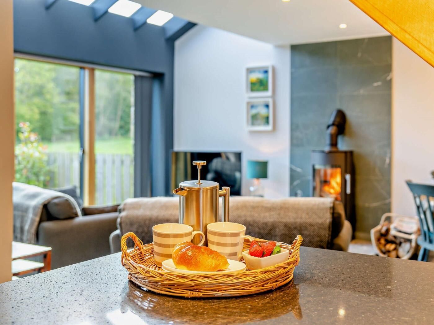 A breakfast setup with a coffee pot, cups, and pastries on a table in a modern living room with a fireplace and large windows.