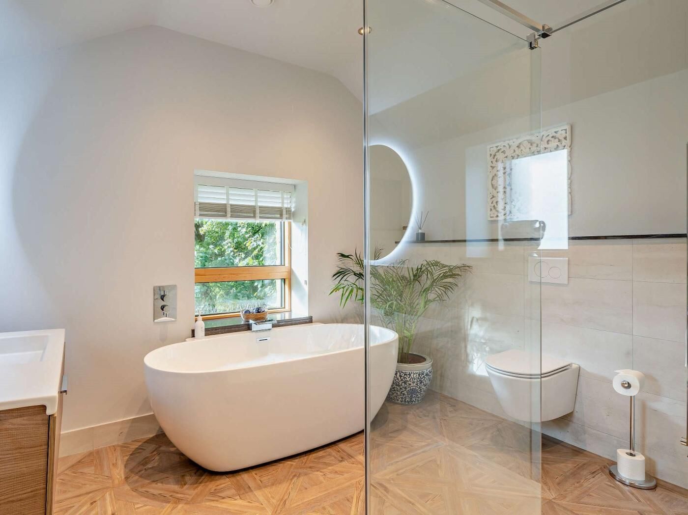 A modern bathroom with a freestanding bathtub, a window, a potted plant, a toilet, and a glass shower enclosure.