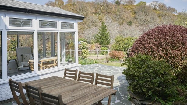 Eine überdachte Terrasse mit Holztisch und -stühlen, ein weißes Gebäude mit großen Fenstern und Blick auf einen bewaldeten Hang.