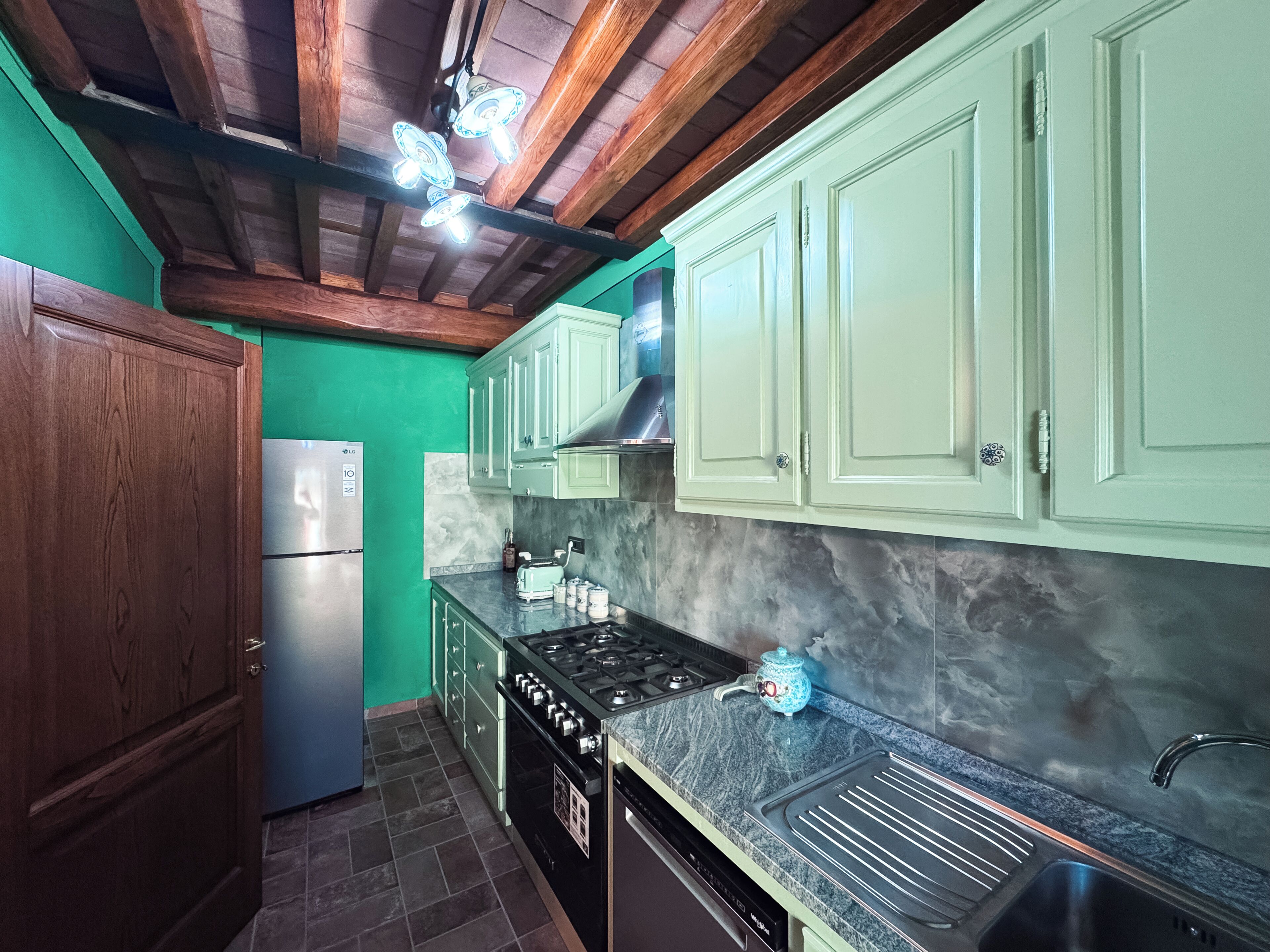A kitchen with wooden beams, green cabinets, a stainless steel refrigerator, a stove, and a sink.