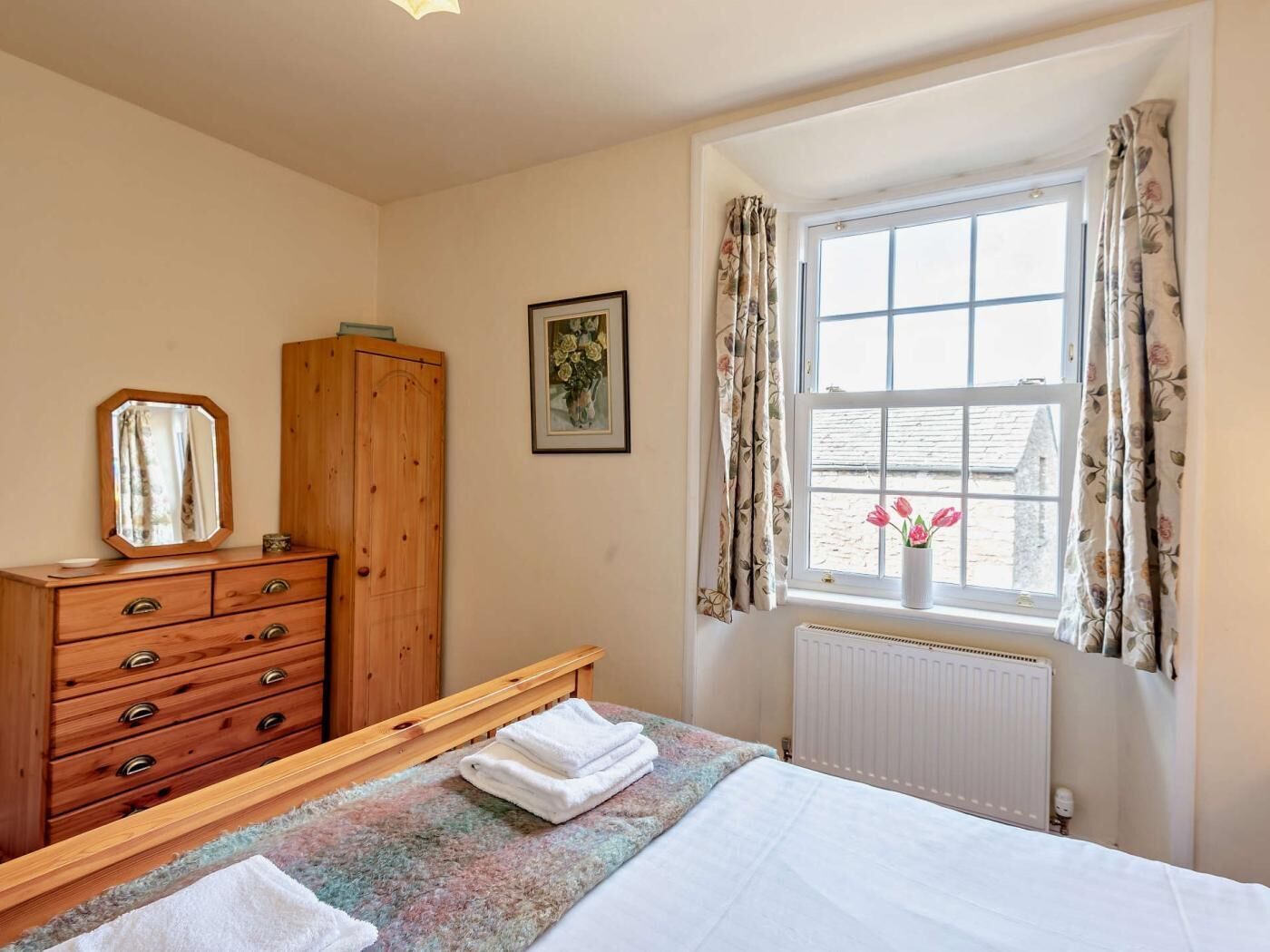 Un dormitorio con cama, cómoda de madera, ventana con cortinas, radiador y un cuadro enmarcado en la pared.