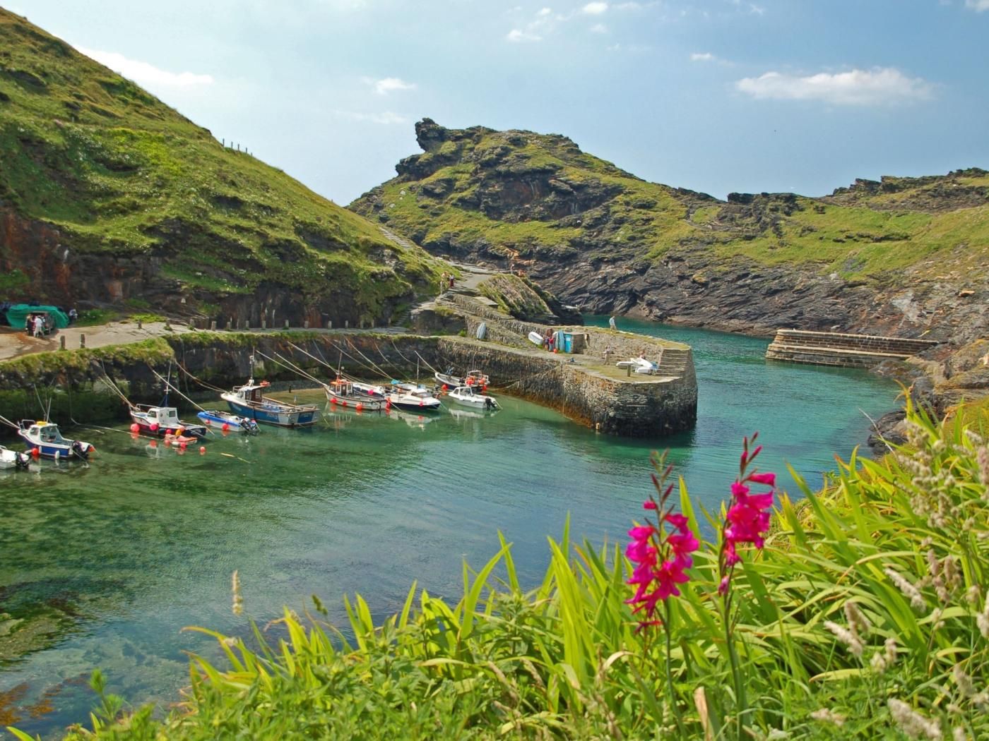 A coastal harbor with boats docked, a stone pier, and a rocky hill in the background.